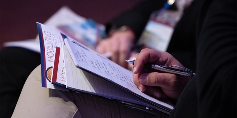 Close up view of symposium flyer and notepad on attendee's lap, the person is holding a pen and taking notes.