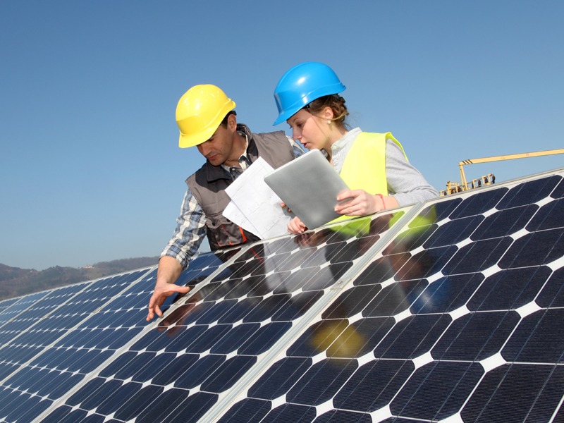 One man and one woman, each with hard hats, leaning over a solar panel.