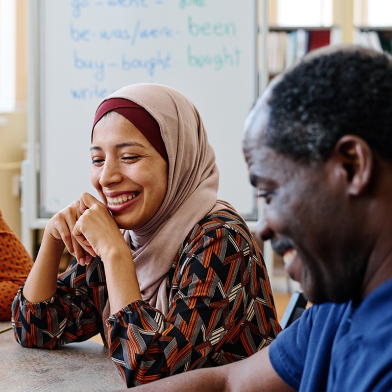 A smiling Muslim woman with her elbows on the table and her hands under her chin and a smiling black man sitting at a table having a discussion.