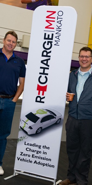 Two men stand by a vertical sign reading Recharge Minnesota, Mankato, Leading the Charge in Zero EMission Vehicle Adoption.