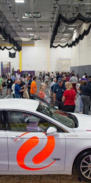 White electric car in foreground, many people at an event talking and looking at exhibits.