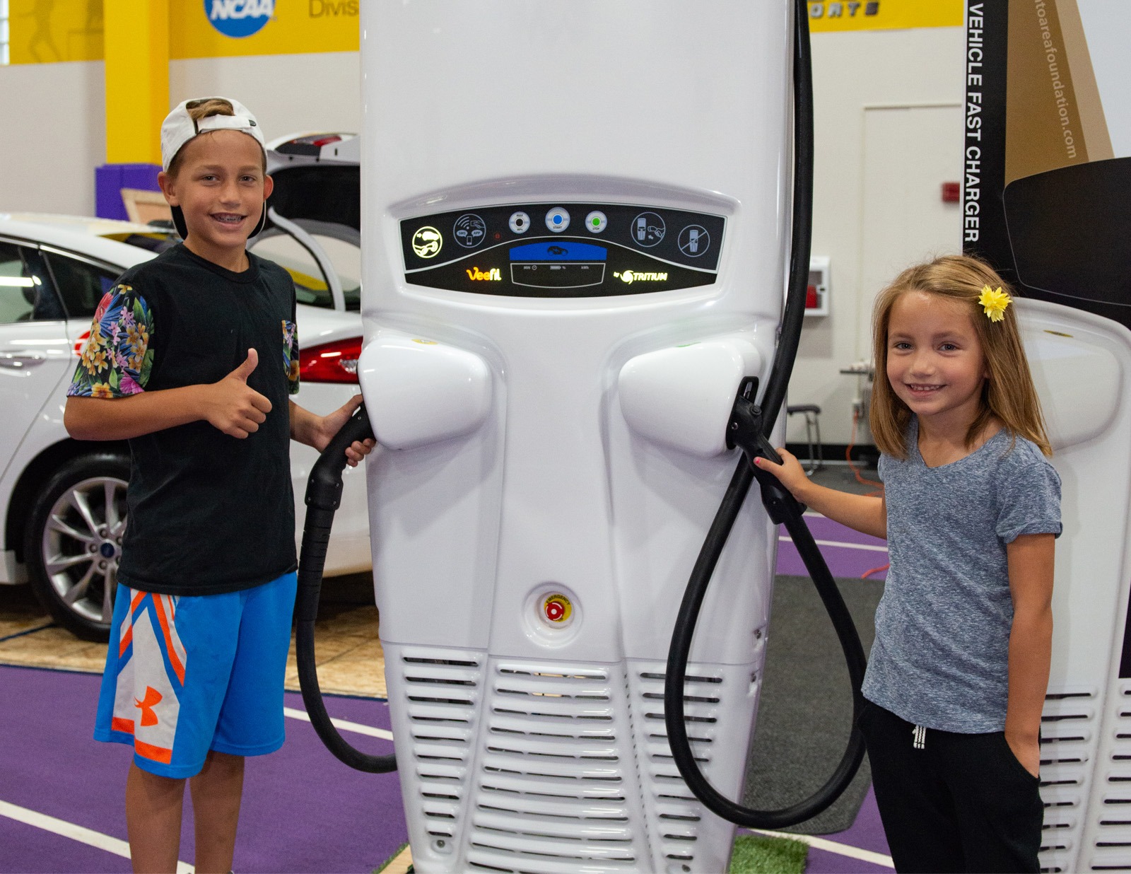 Two smiling young children standing by an electric vehicle charger.